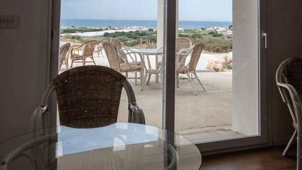 Een kamer met een glazen tafel en rieten stoelen, met uitzicht op een strand en een helder uitzicht over de oceaan en gebouwen in de verte.