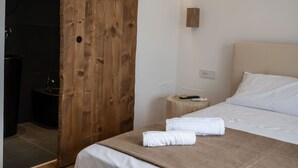 Een hotelkamer met een houten deur, een bed met witte lakens, een nachtkastje met een afstandsbediening en een klein rond nachtkastje.