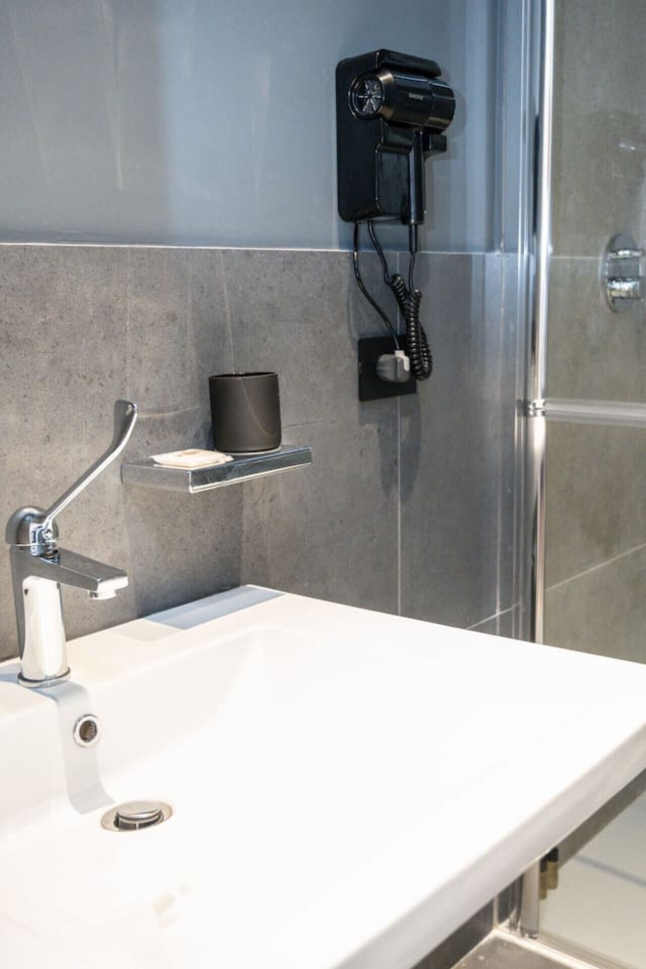 A modern bathroom with a white sink, a handheld showerhead, a towel rack, and a hairdryer mounted on the wall.