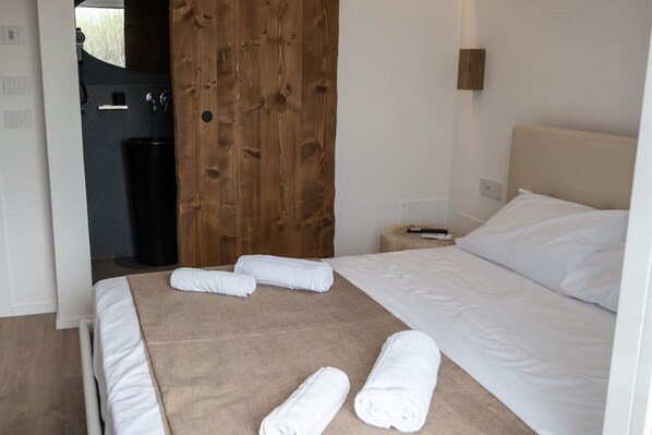 Habitación de hotel con cama, puerta de madera, cubo de basura negro y una mesita de noche con control remoto.