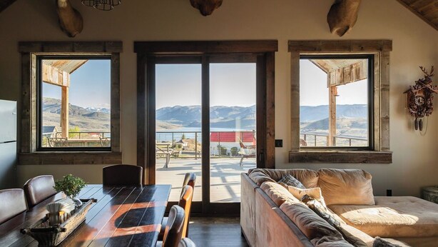 A spacious room with a wooden dining table, a sofa, and large windows offering a view of mountains and a lake. - Modern Cabin at Blue Mesa, INCREDIBLE VIEWS, 40min from BLK CNYN (Gunnison)