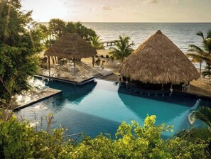 Zona de piscina en un resort con dos estructuras de techo de paja, vista despejada del océano y abundante vegetación.