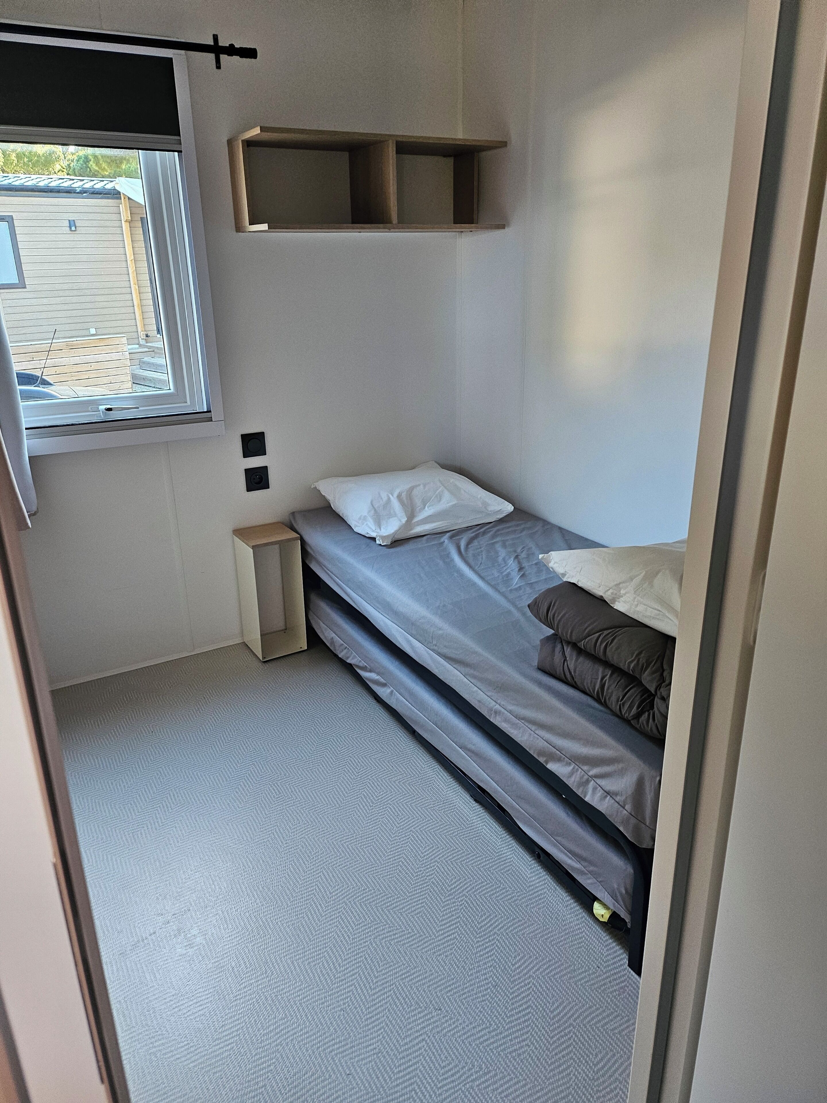A small, compact room with a single bed, a window, and a small shelf.