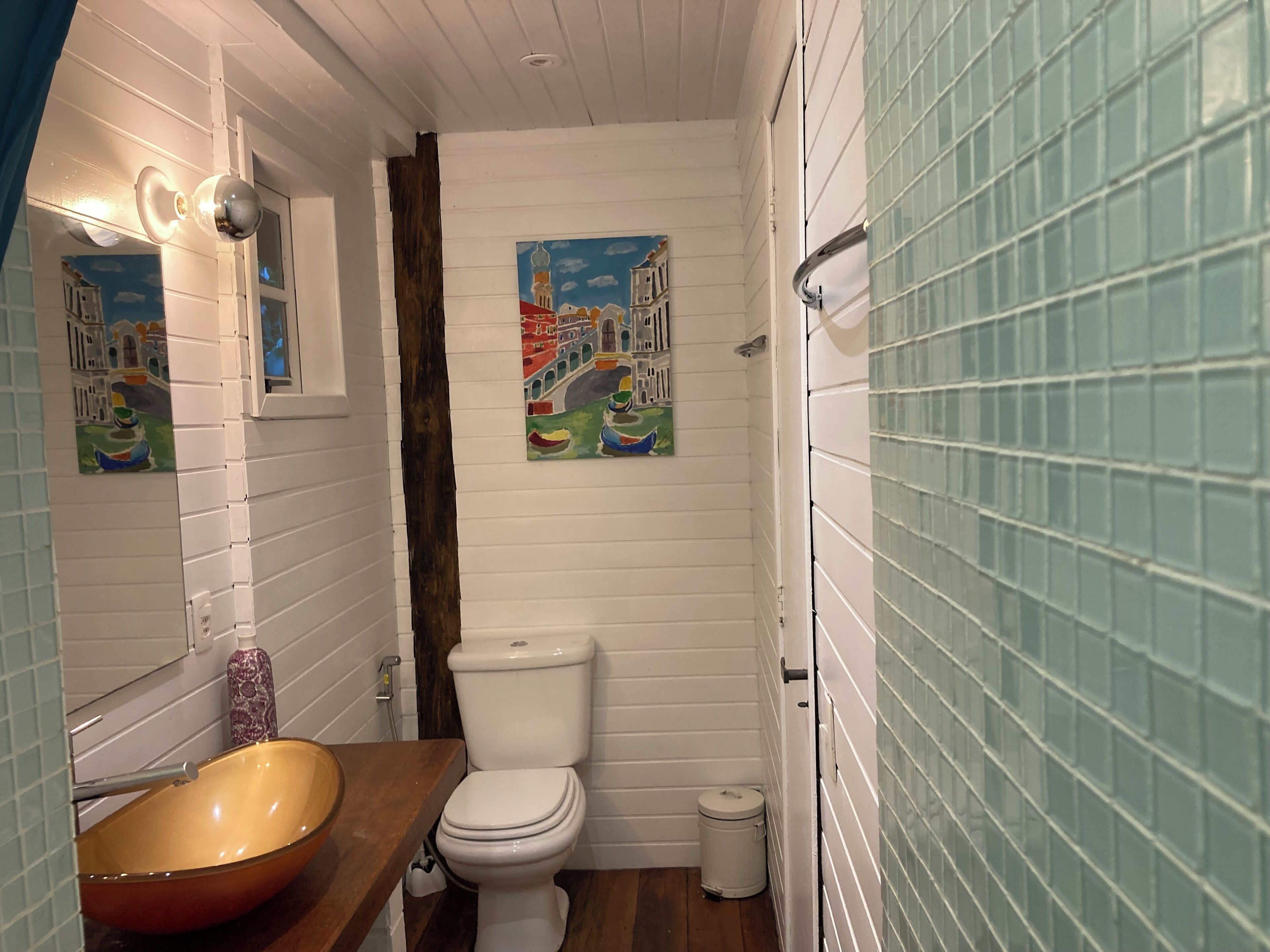 Un baño con tocador de madera, inodoro blanco, un pequeño cubo de basura y un cuadro con vista urbana en la pared.