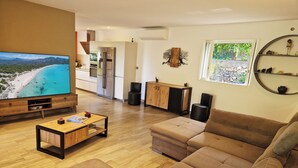 A modern living room with a large flat-screen TV, a wooden coffee table, a brown sofa, and a kitchen in the background.