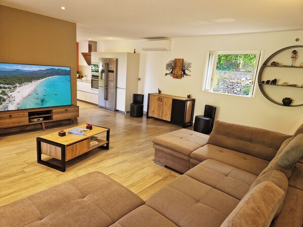 Una sala de estar moderna con un televisor de pantalla plana grande, una mesa de centro de madera, un sofá marrón y una cocina al fondo.