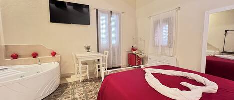 A hotel room with a large bed, a small table with chairs, a desk, a bathtub, and a window with curtains.