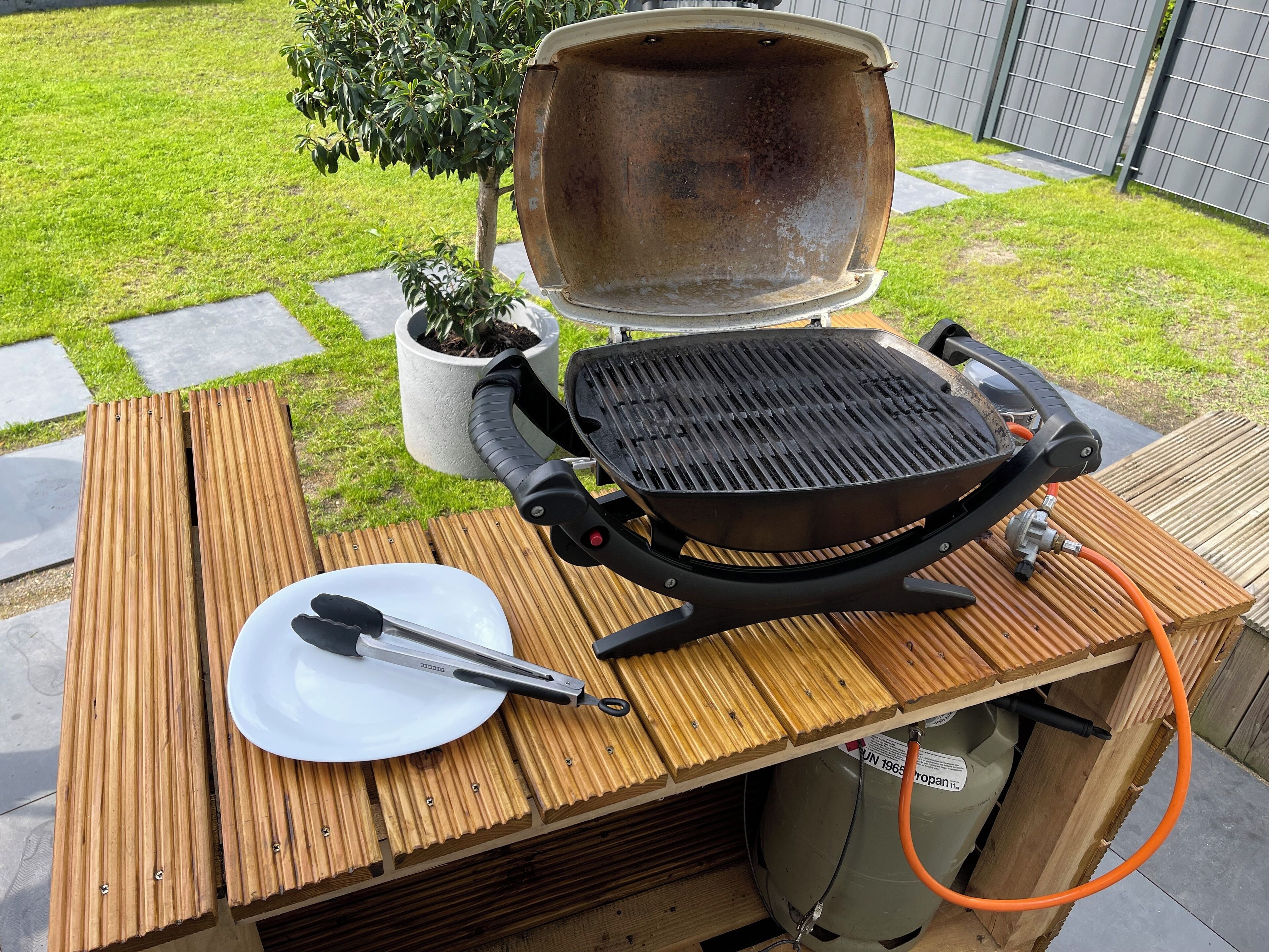 Ein tragbarer Grill auf einem Holztisch mit einem weißen Teller und Zangen, verbunden mit einer Propangasflasche.