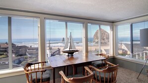 A room with a large window overlooking a beach and ocean, a wooden table with chairs, and a sailboat model on the table. - Haystack Haven-Haystack Rock View-Sunset Views-Popular Destination-Unwind (Cannon Beach)