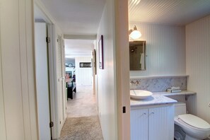 Un couloir étroit mène à une salle de bain équipée d’un lavabo, d’un miroir et de toilettes.