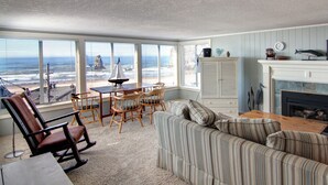 A living room with a view of the ocean, a fireplace, and a dining area with a sailboat model. - Haystack Haven-Haystack Rock View-Sunset Views-Popular Destination-Unwind (Cannon Beach)