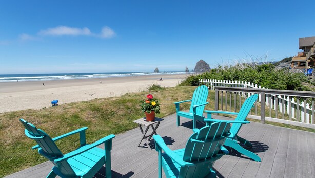 Une terrasse dotée de chaises Adirondack turquoise, d’une petite table ornée d’une plante en pot et offrant une vue sur la plage et l’océan.