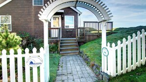 Une maison en bois avec un porche, une clôture blanche en piquets et un chemin de pierre menant à l’entrée.
