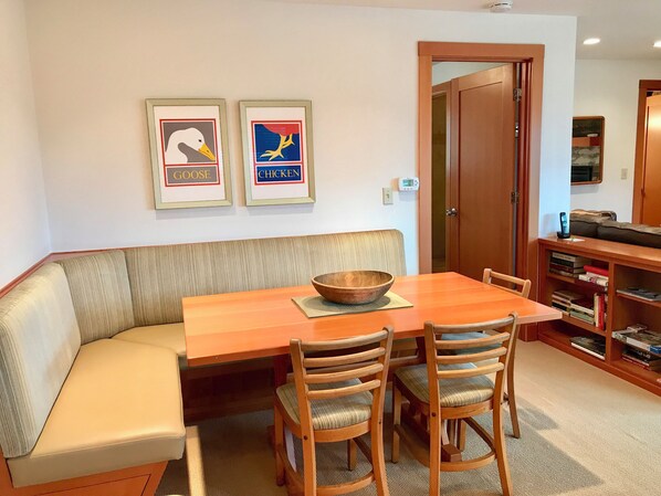 En matplats med ett träbord, fyra stolar, en träskål och en bänk. På väggen hänger två inramade tavlor, varav den ena är märkt med texten "GOOSE" och den andra med "CHICKEN". I bakgrunden syns en bokhylla med böcker.