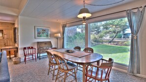 A dining area with a wooden table and chairs, a painting on the wall, and a view through large windows.