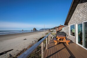 Une terrasse en bois avec une table de pique-nique et un barbecue, donnant sur une plage avec un promontoire rocheux en arrière-plan.