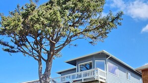 Ein zweistöckiges Haus mit Balkon, ein Baum im Vorgarten und ein klarer, blauer Himmel.
