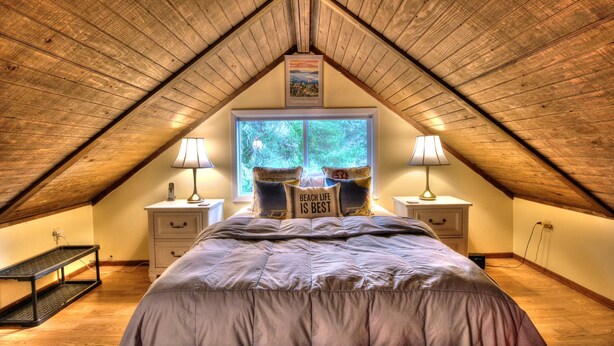 Une chambre mansardée chaleureuse avec un plafond en bois incliné, un grand lit donnant sur la verdure, deux tables de chevet avec des lampes et un tableau encadré au-dessus de la fenêtre.