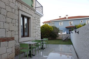 A patio with green chairs and a table, a stone wall, and a blue building in the background. - Cemile Hanim Konagi (Cesme)