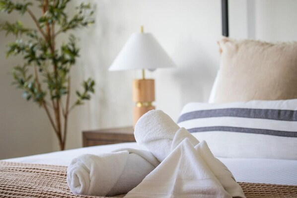 A neatly made bed with white bedding and a striped pillow, a white towel on the bed, a wooden bedside table with a lamp, and a plant in the background. - The Wright House - Modern 3BDRM home in Midland! (Midland)