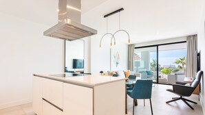 A modern kitchen with a central island, a dining area with a blue chair, and a view of the sea through large windows. - Santa Barbara Heights IV CLC - Luxury, private plunge pool (Mijas Costa)