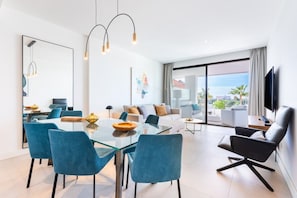 Een moderne woonkamer met een glazen eettafel, blauwe stoelen, een zwarte leren bank en een flatscreen televisie.