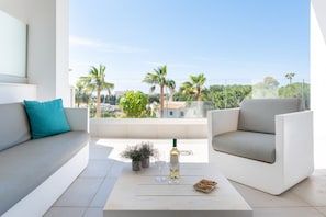 Un área de estar moderna al aire libre con un sofá y sillones, una mesa de centro con una botella y vasos, y vistas a palmeras y edificios.