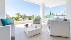 A modern living room with white furniture, a large window offering a view of palm trees and a pool, and a small kitchen area. - Santa Barbara Heights IV CLC - Luxury, private plunge pool (Mijas Costa)