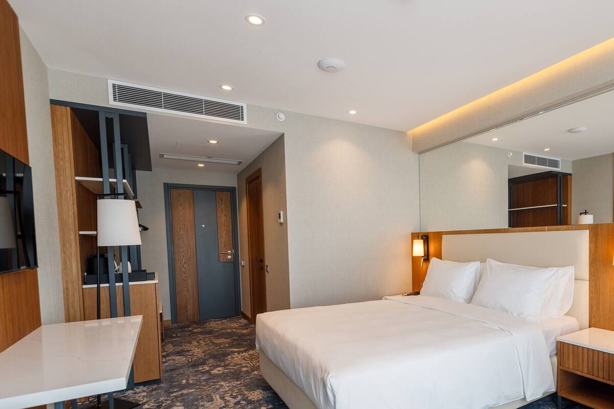 Une chambre d’hôtel moderne avec un grand lit, un bureau et une chaise. Un miroir est placé au-dessus du lit et une petite table se trouve à côté. La chambre est dotée d’un sol recouvert de moquette et d’une porte qui donne sur une autre pièce.
