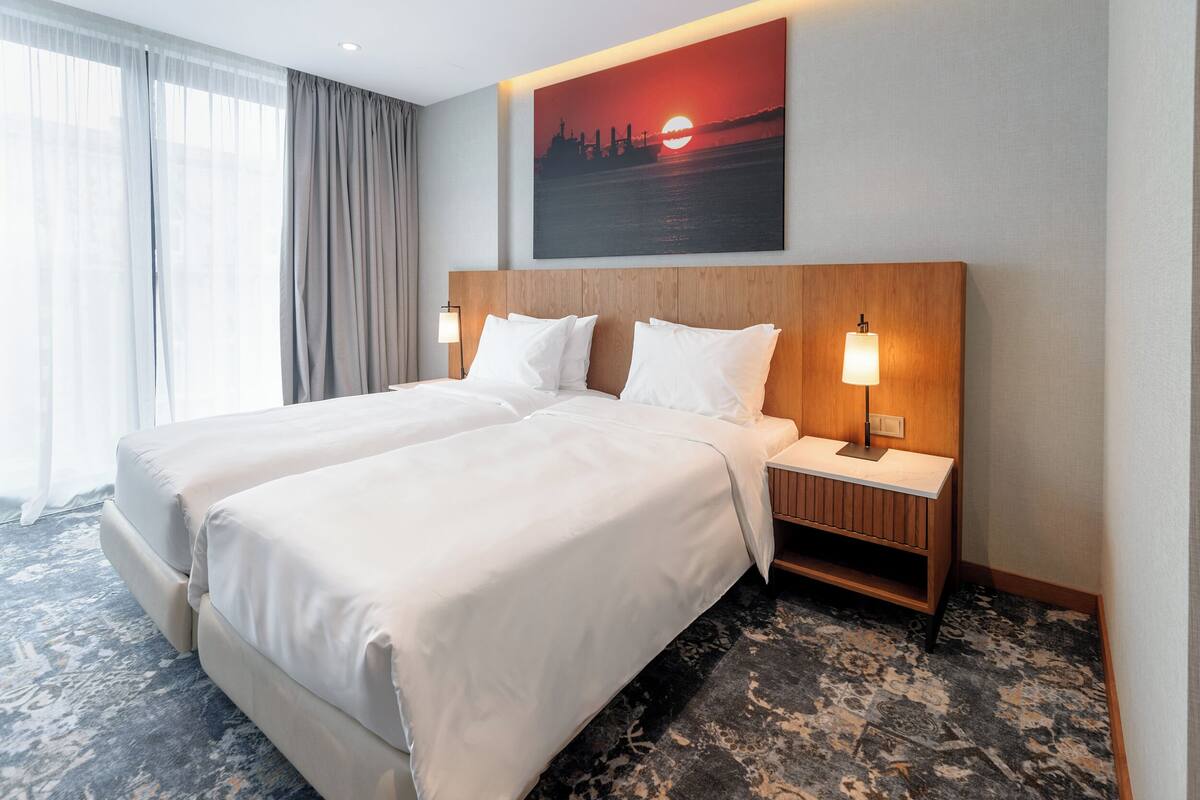 Une chambre d’hôtel avec deux lits, une table de chevet avec une lampe, un tableau représentant un coucher de soleil et une grande fenêtre avec des rideaux.