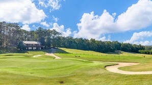 Un campo de golf con un club de campo, búnkeres de arena y un área boscosa al fondo.