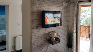 A room with a brick wall, a mounted television, a radiator, and a window with a view of a wooden deck.