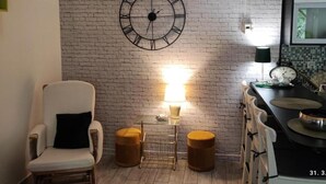 A room with a brick wall, a large clock, a chair, a small table, and a lamp.