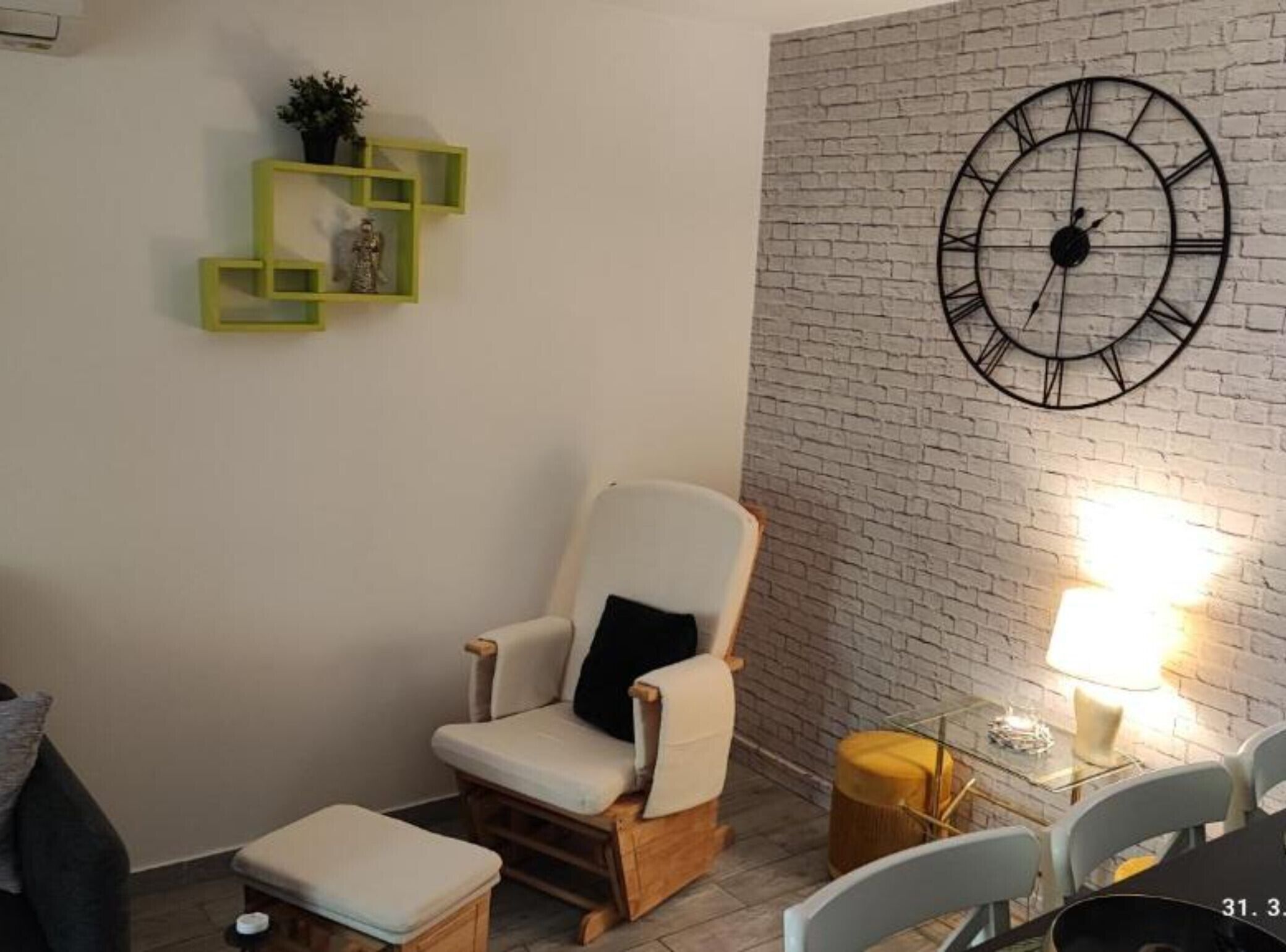 Un rincón acogedor con un sillón mullido, un pequeño taburete, una mesa redonda con una lámpara y un reloj decorativo en una pared de ladrillo.