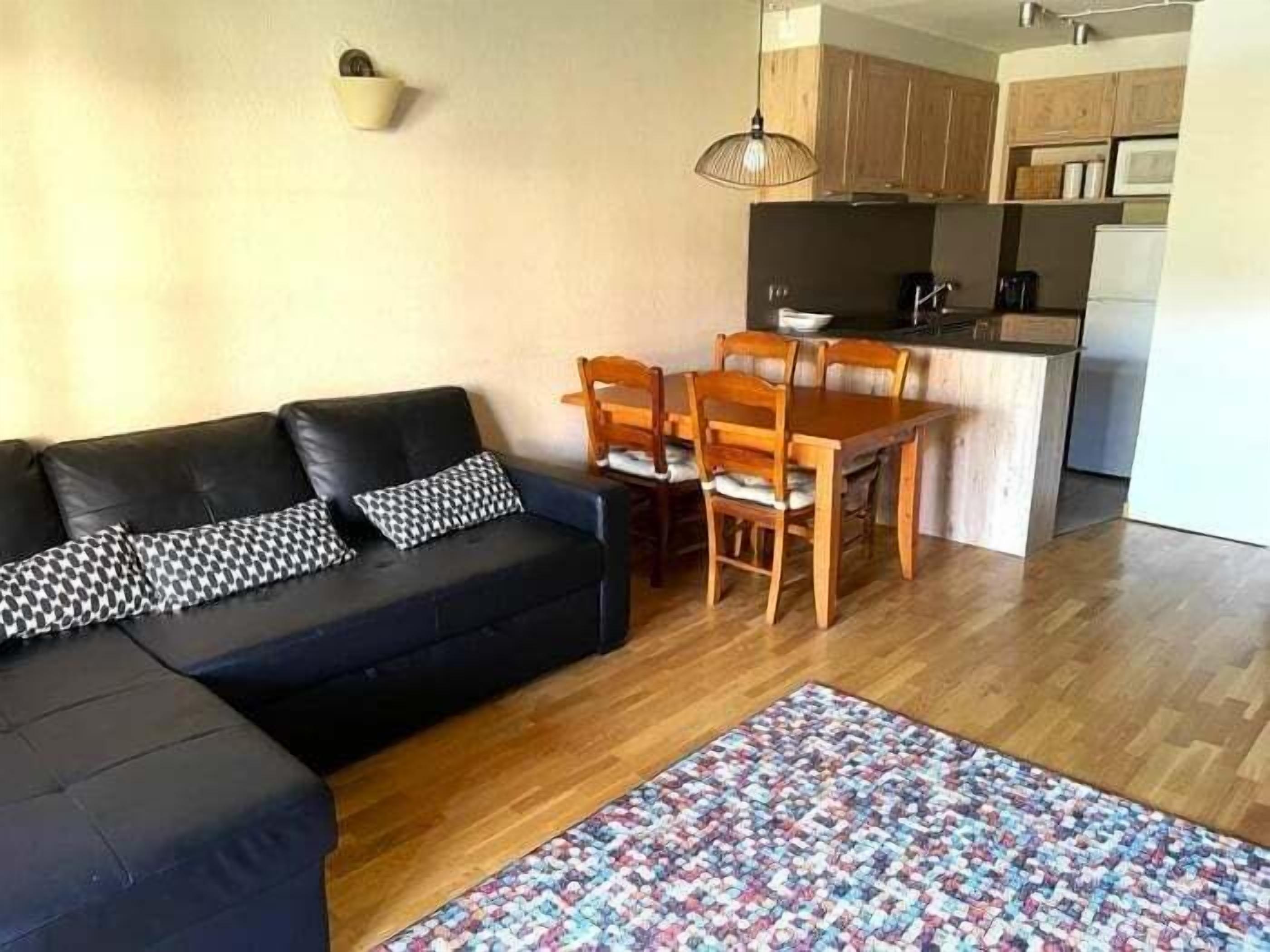 Un soggiorno con un divano nero, cuscini decorati, un tavolo da pranzo in legno con sedie, angolo cottura con mobili in legno e un tappeto a motivi.