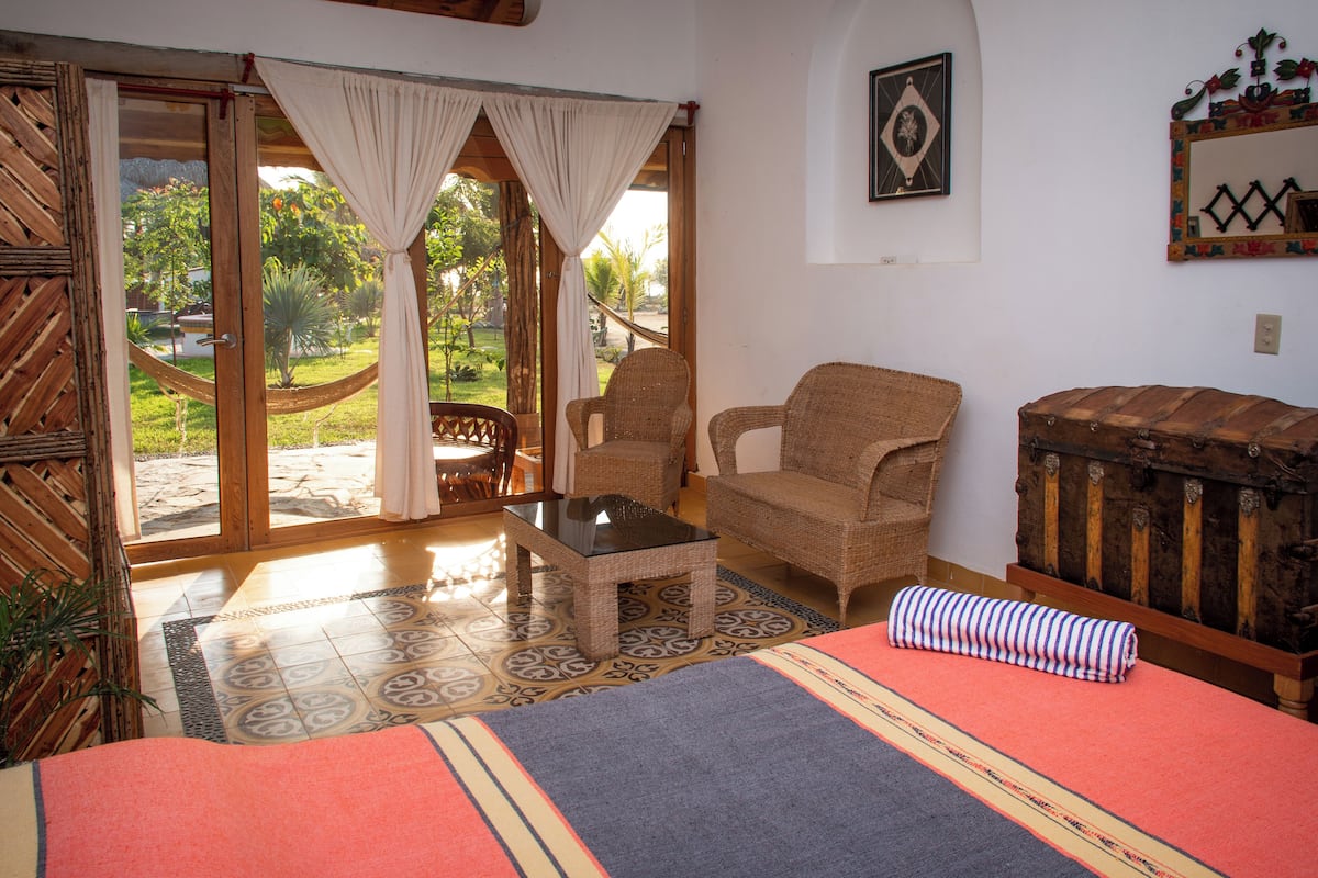 A room with a bed, a wicker chair, a small table, and a view of a garden through a glass door.