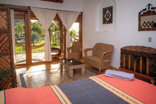 A room with a bed, a wicker chair, a small table, and a view of a garden through a glass door. - El MURO MAJAHUA RETREAT CENTER (Troncones)