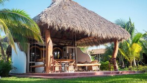 A thatched-roof structure with a dining area and outdoor seating, surrounded by palm trees and greenery. - El MURO MAJAHUA RETREAT CENTER (Troncones)
