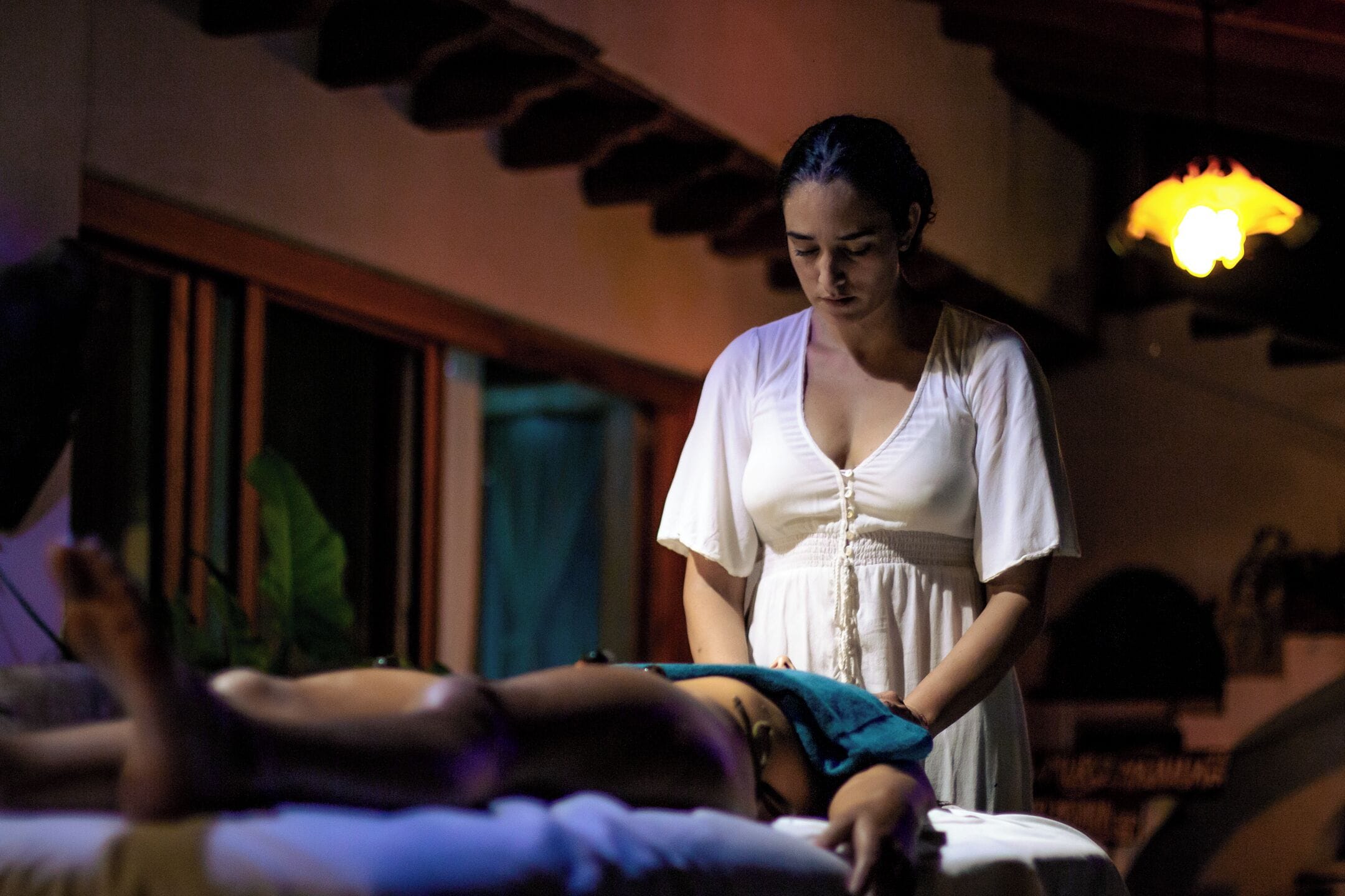 A person lying on a bed in a dimly lit room with a window and a hanging light fixture.