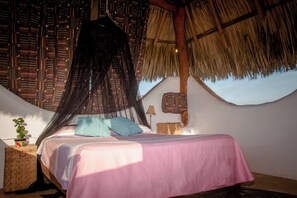 A bed with a pink bedspread, a blue pillow, and a mosquito net. There is a small table with a plant and a lamp. The room has a thatched roof and patterned walls. - El MURO MAJAHUA RETREAT CENTER (Troncones)