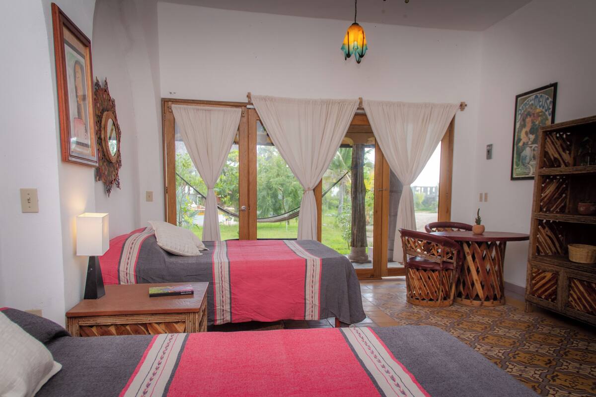 A room with two beds, a table, chairs, a hammock, and a view of greenery outside.