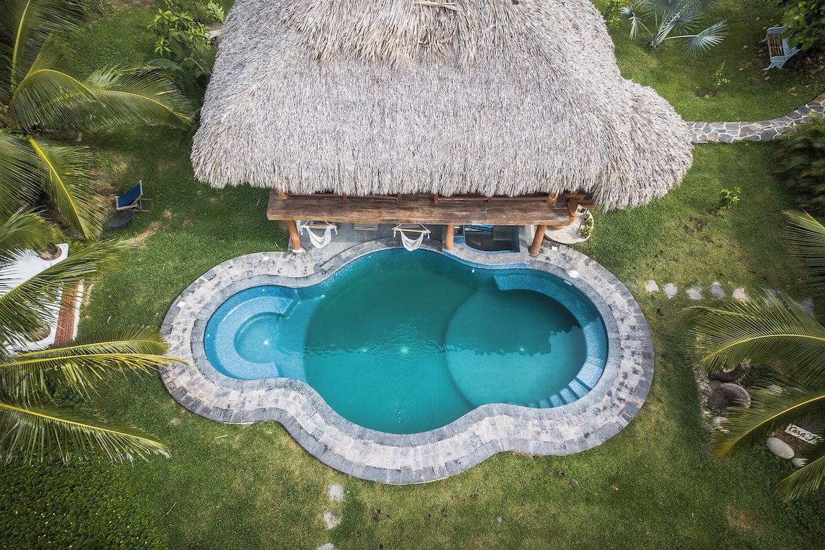 A thatched-roof structure with a circular pool, surrounded by palm trees and a stone pathway.