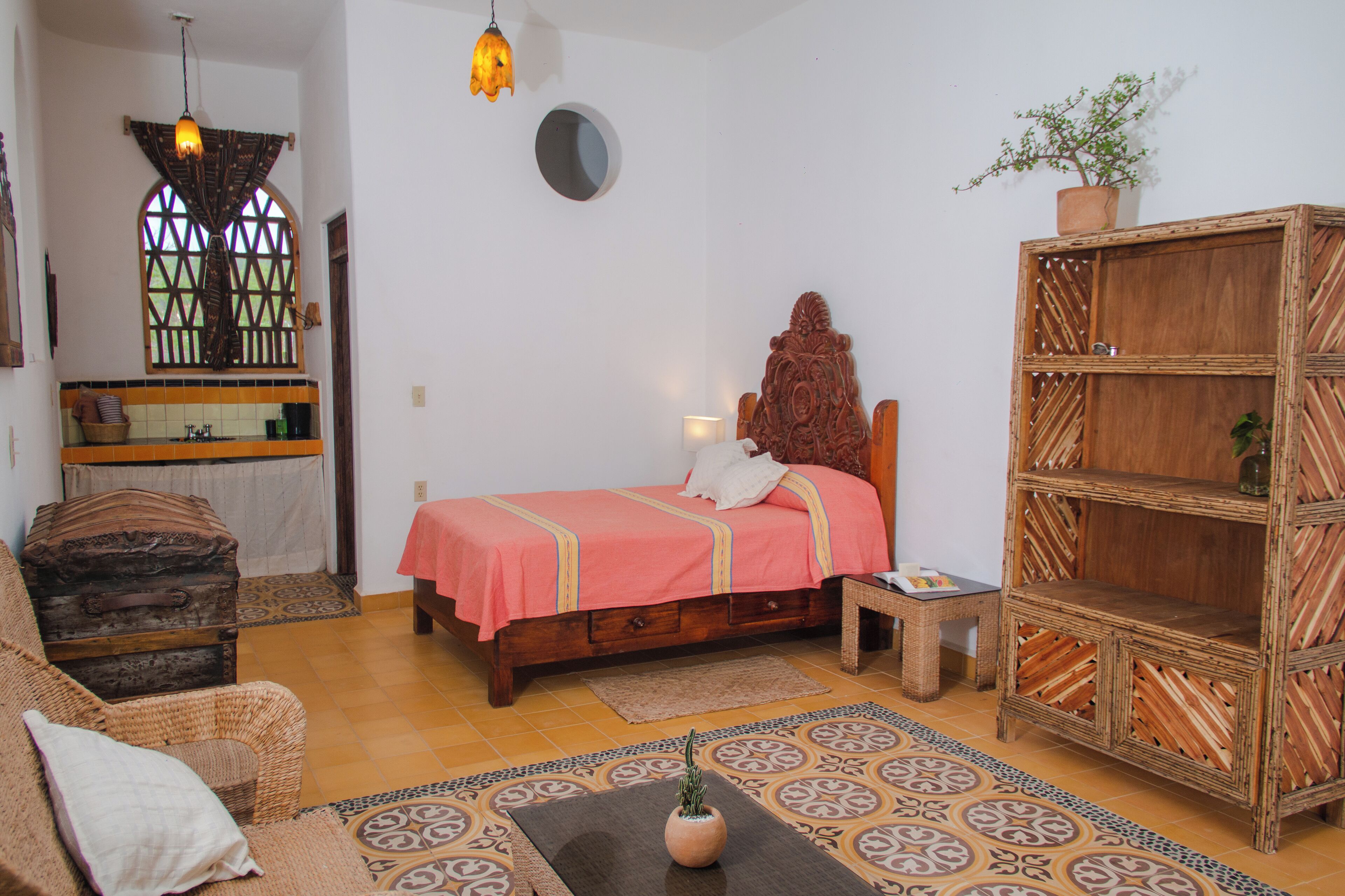 A bedroom with a bed, a wooden headboard, a wicker chair, a small table, a shelf, and a potted plant.