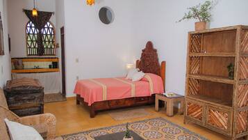A bedroom with a bed, a wooden headboard, a wicker chair, a small table, a shelf, and a potted plant.