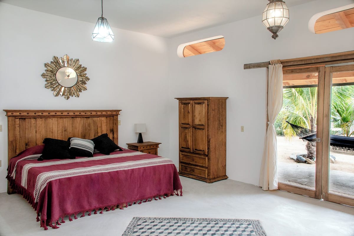 A bedroom with a wooden bed, a nightstand, a wardrobe, and a view of a tropical garden through a sliding door.