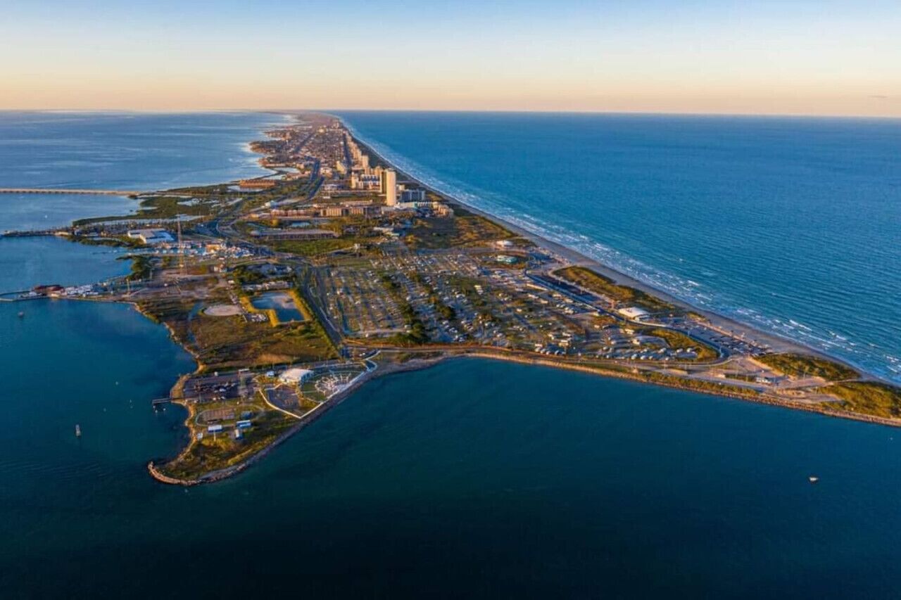 Vista aérea de una ciudad costera con una extensa playa, una zona urbana densa y una península con edificios industriales y residenciales.