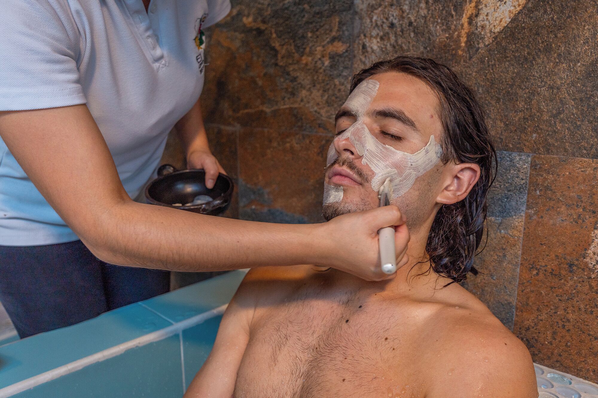 Una persona recibiendo un tratamiento facial en un baño con pared de piedra y lavamanos con azulejos azules.