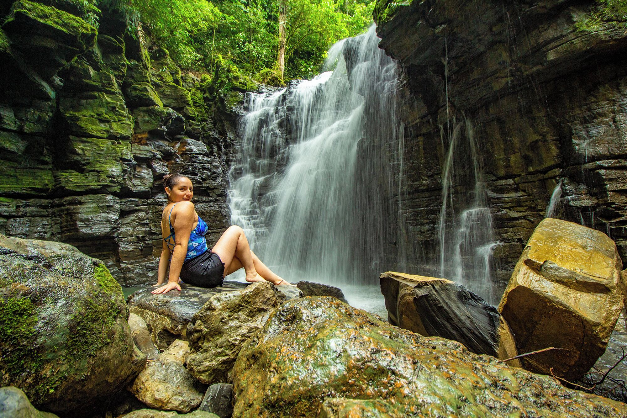Una persona sentada sobre rocas cerca de una cascada en una zona boscosa.