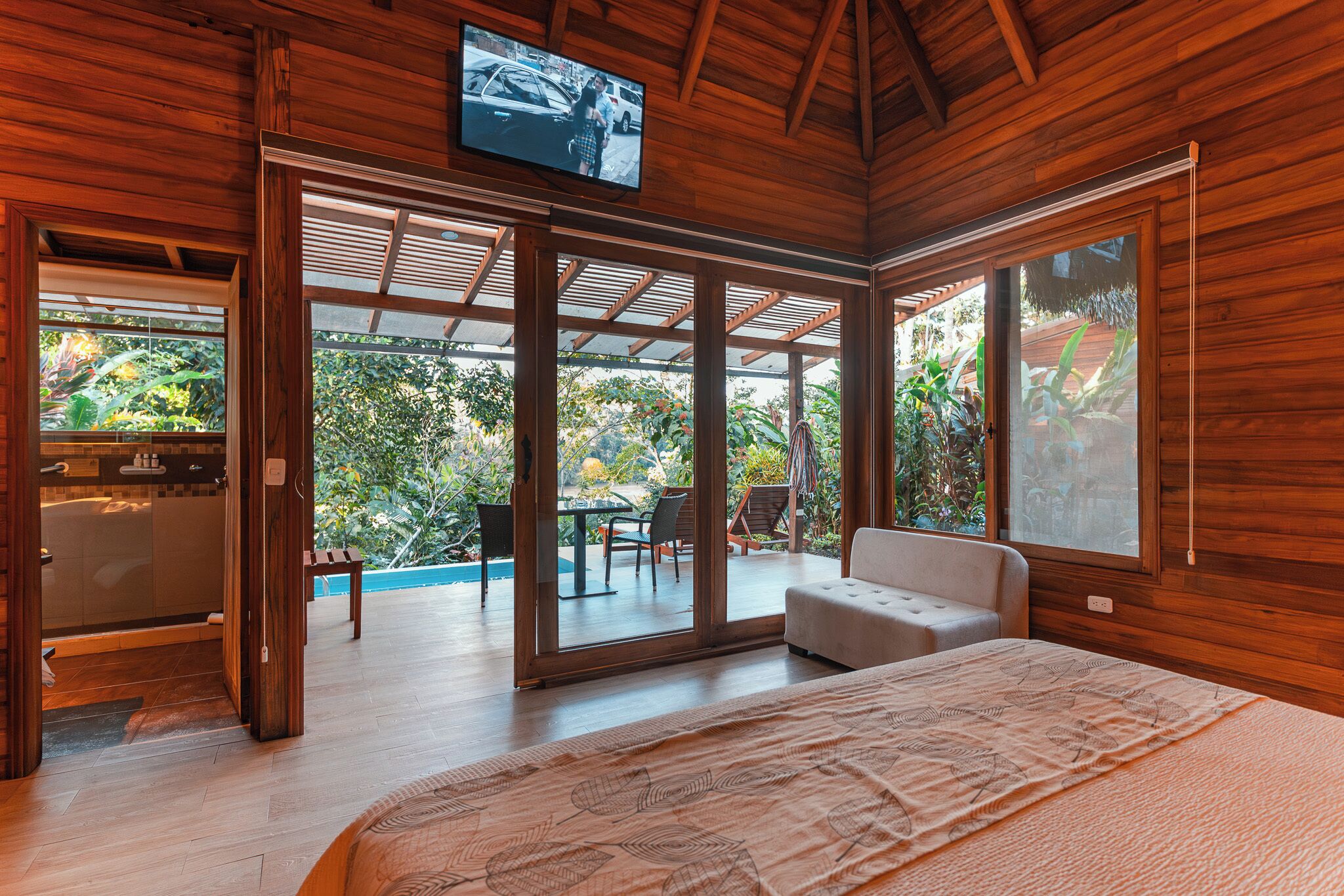 A spacious bedroom with a large bed, sliding glass doors leading to a patio with outdoor furniture, and a television mounted on the ceiling.