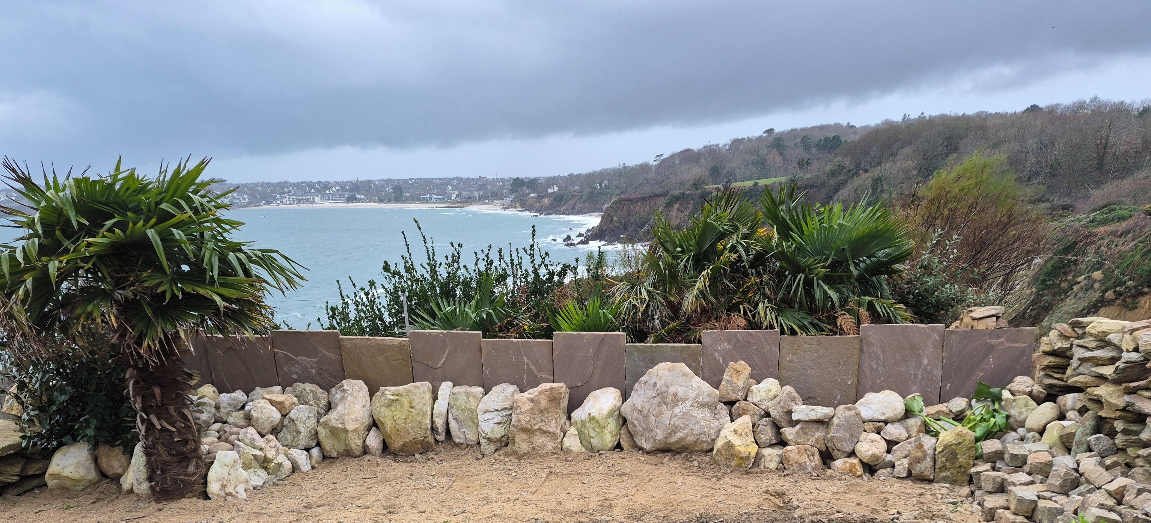 A coastal landscape with a rocky barrier, vegetation, and a view of the sea and distant buildings.